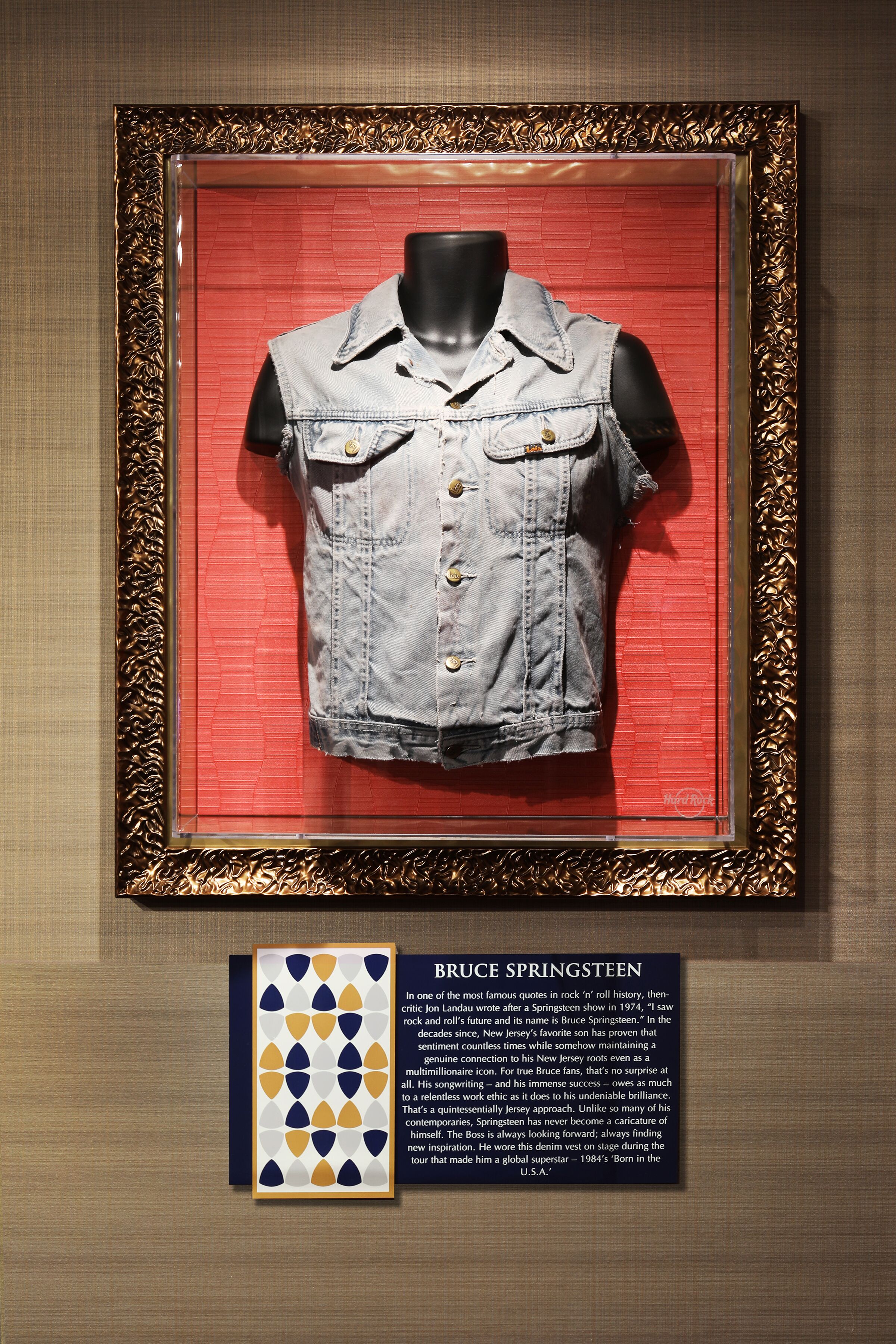 Uma jaqueta jeans exibida em uma vitrine de vidro com moldura dourada e fundo vermelho. Abaixo da jaqueta, há um painel com moldura que apresenta o nome 'Bruce Springsteen' e uma citação.