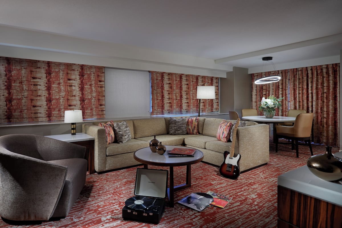 A modern hotel room with a sofa, armchair, dining table, and a guitar.