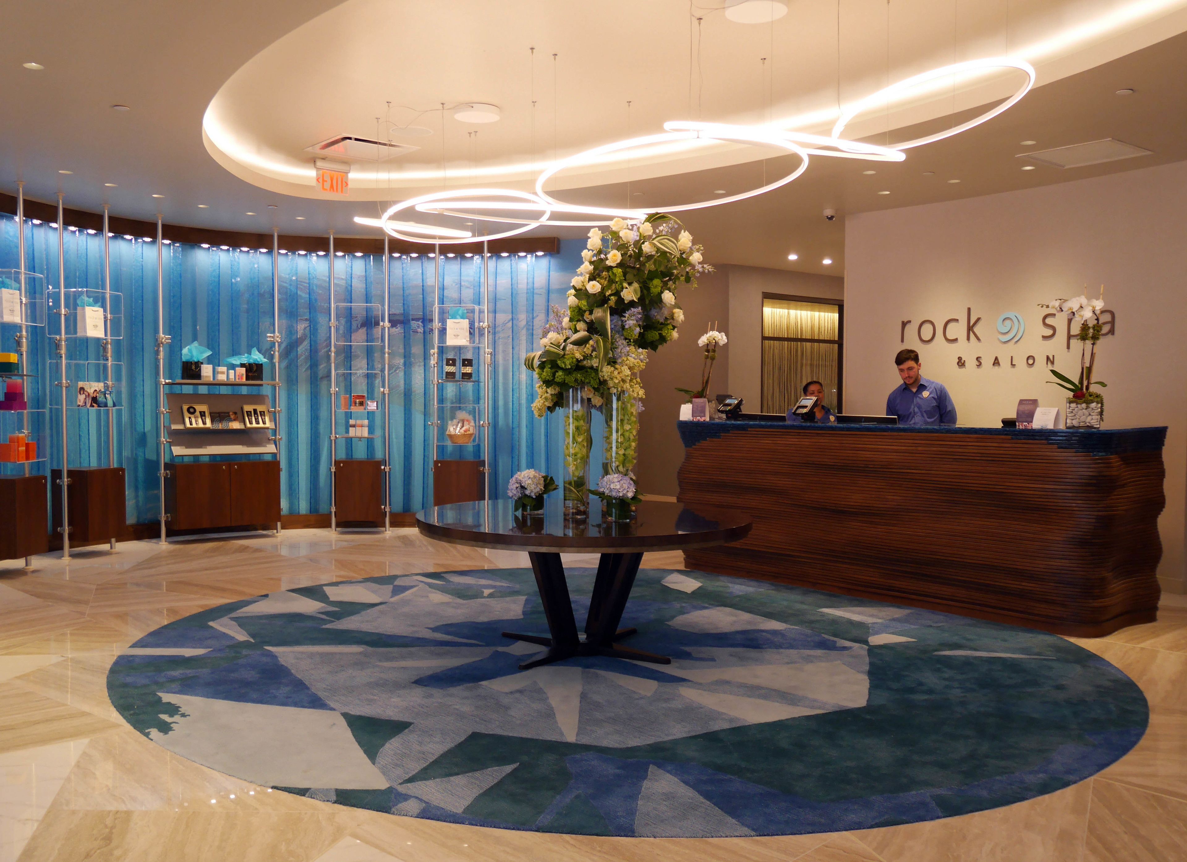 A reception area with a modern design, featuring a central round table with a floral arrangement, a reception desk, and a wall with a sign reading "rock spa & salon".