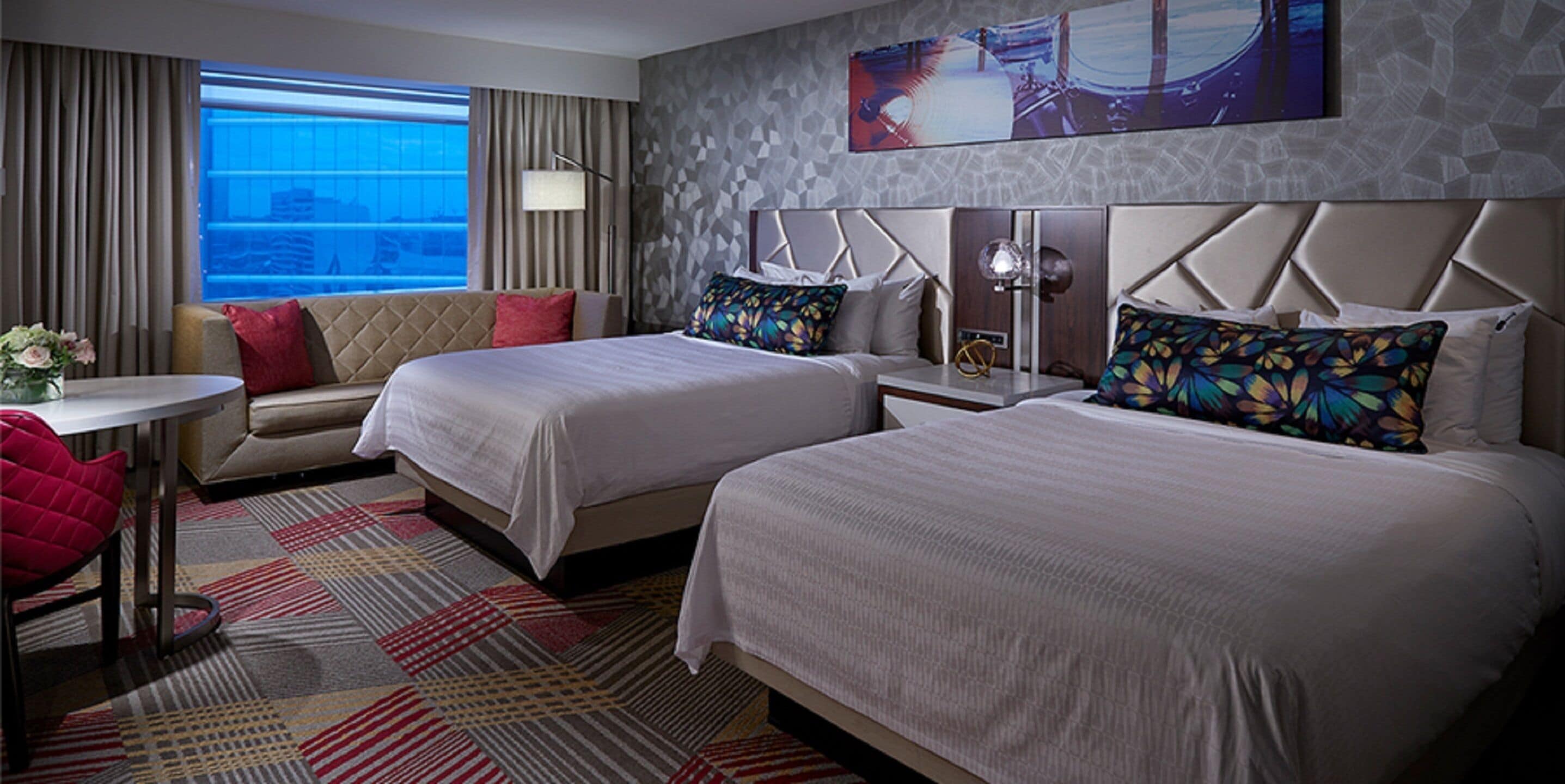 A hotel room with two beds, a sofa, a desk, and a large window offering a city view.