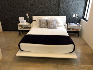 A modern bedroom with a white bed, two decorative pillows, a dark throw blanket, and a nightstand with a lamp and books.