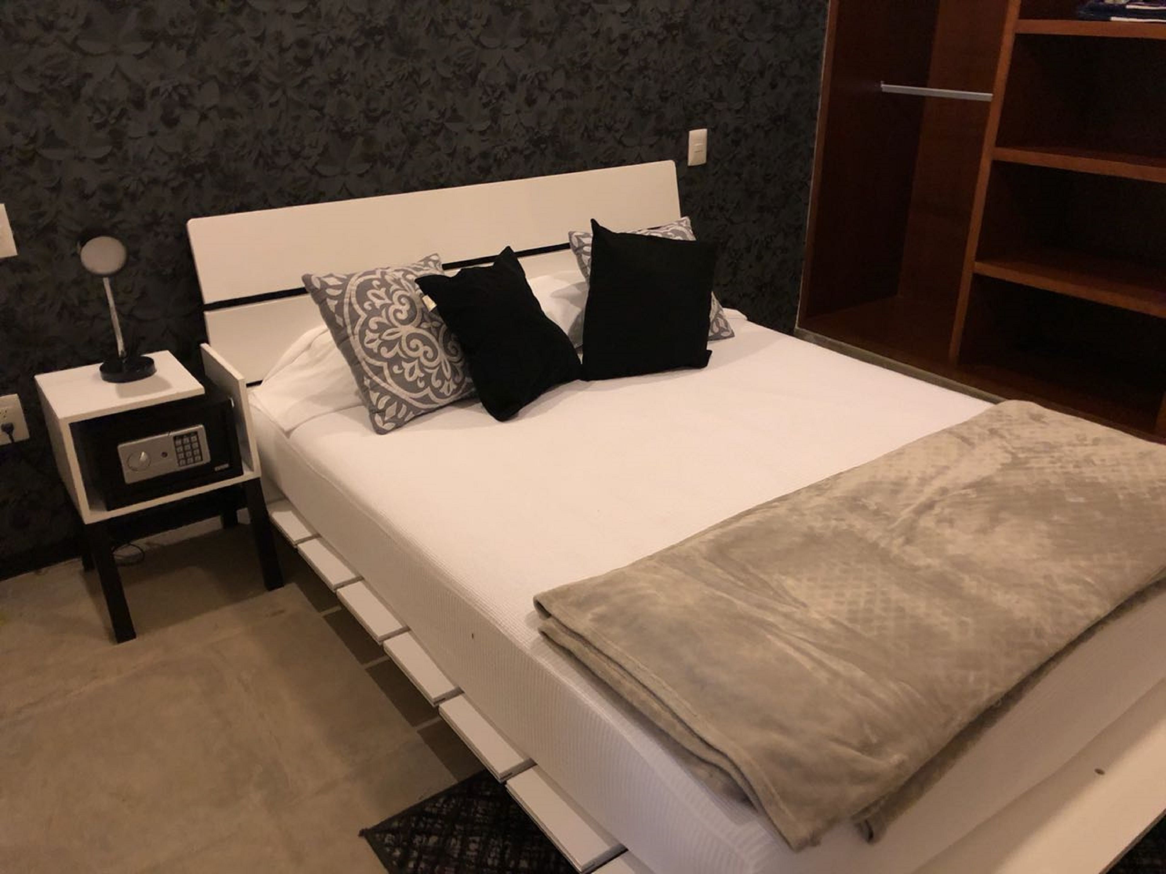 Ein ordentlich bezogenes Bett mit einer beigen Decke, einem weißen Kopfteil und zwei schwarzen sowie einem weißen Kopfkissen. Ein kleiner Nachttisch mit einer Lampe und einer Steckdose steht daneben.
