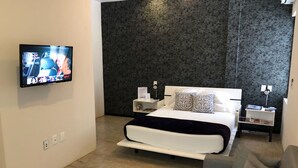 A modern bedroom with a large bed, a TV mounted on the wall, a bedside table with a lamp, and a small side table with books. - Downtown Suites Cancun (Cancun)