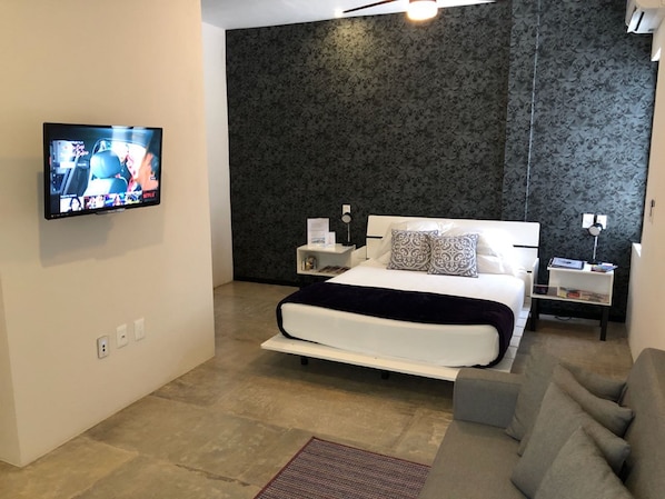 A modern bedroom with a large bed, a TV mounted on the wall, a bedside table with a lamp, and a small side table with books.
