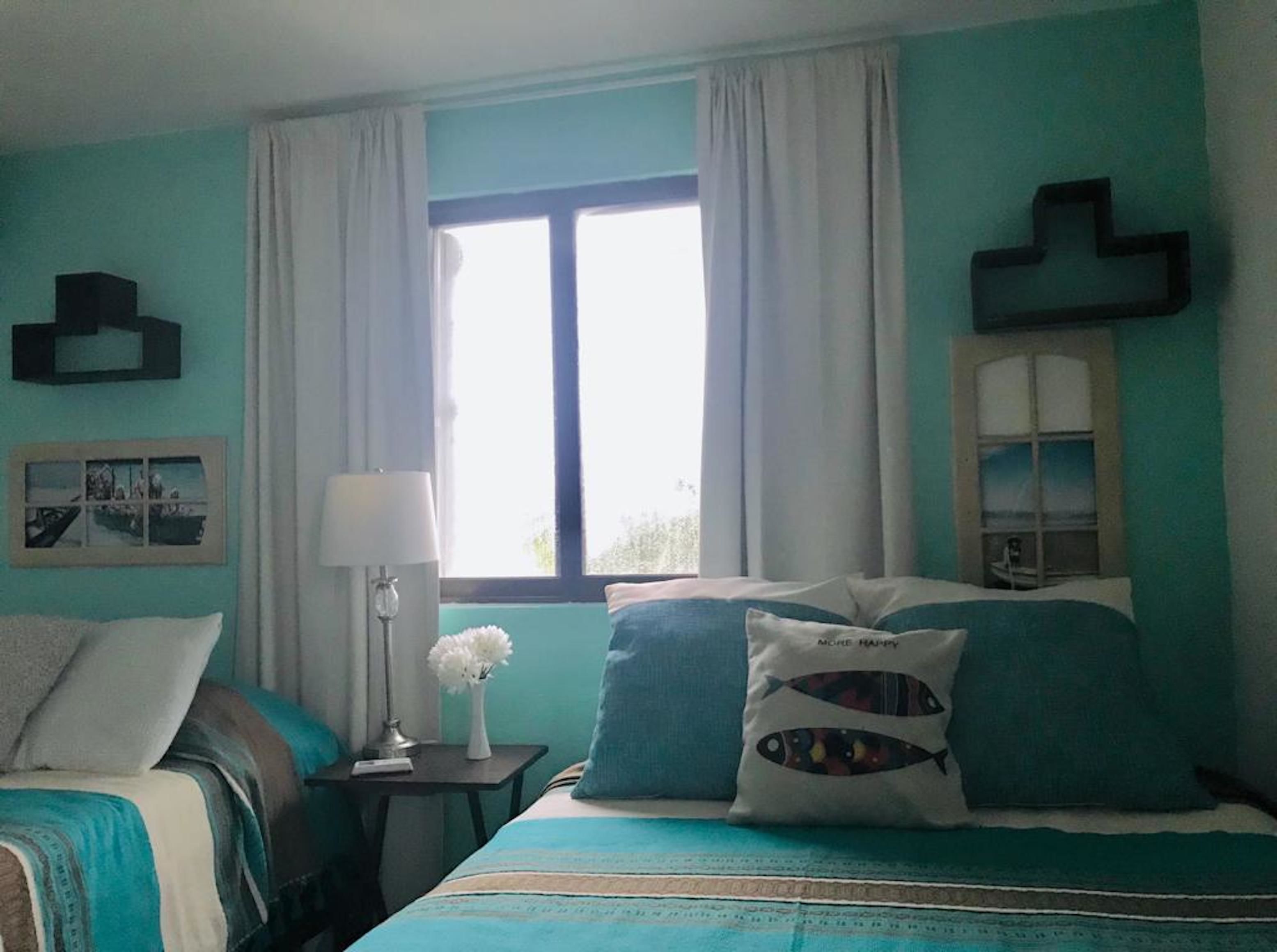 A bedroom with a bed, a nightstand, a lamp, a window with curtains, and a wall-mounted mirror.
