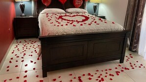 Een slaapkamer met een bed, een rozenblaadjes-decoratie in de vorm van een hart, rode hartvormige ballonnen, en een bordje met de tekst 'liefde' aan de muur.