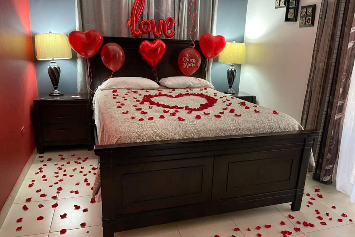 Une chambre avec un lit, une décoration en forme de cœur faite de pétales de roses, des ballons rouges en forme de cœur et un panneau avec le mot « amour » accroché au mur.