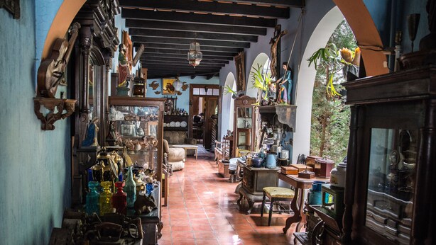 A narrow corridor with antique furniture and various items on display, leading to a door at the end. - Posada San Sebastian (Antigua Guatemala)