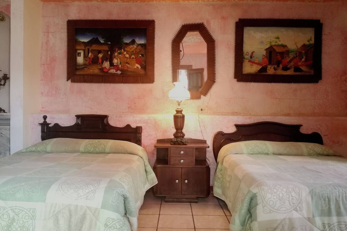 A bedroom with two beds, a nightstand with a lamp, and two wall-mounted paintings.