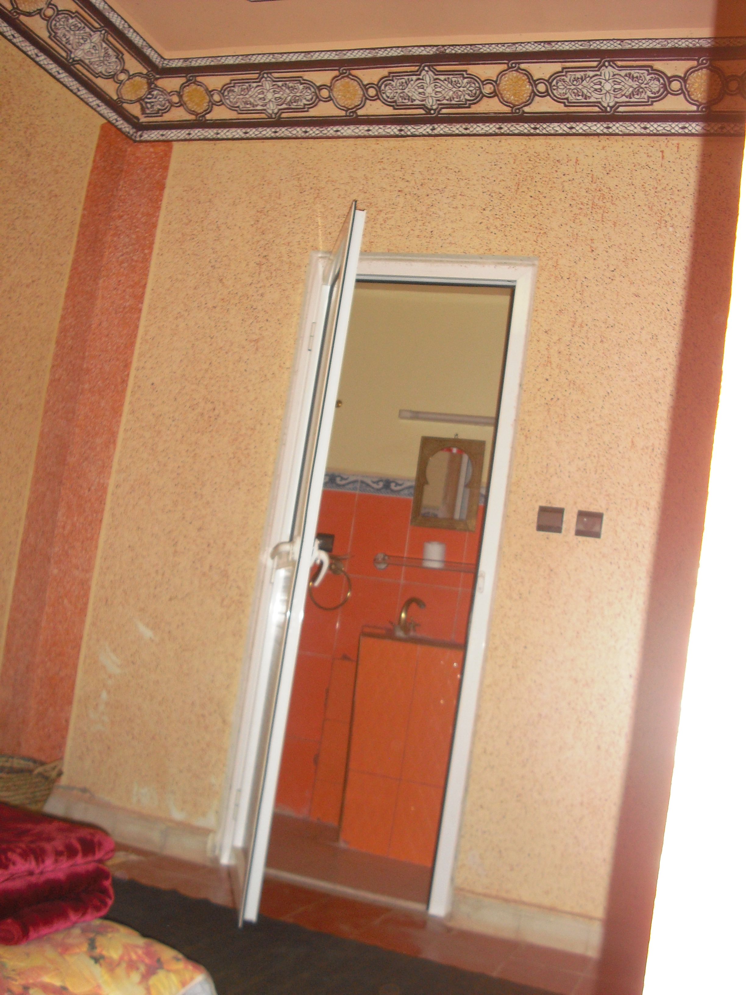 A room with a door leading to a bathroom. The bathroom has a sink, mirror, and towel rack. The room has a patterned wall and a red carpet.