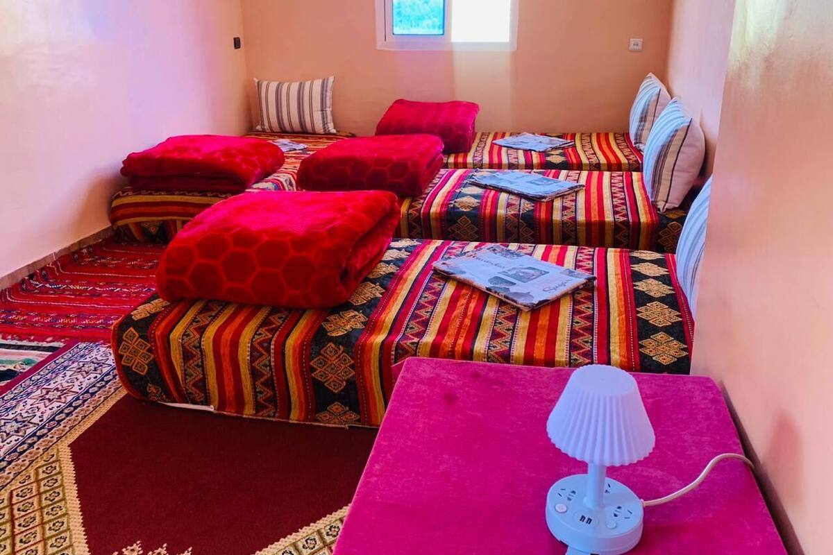 Una habitación con dos camas, cada una con ropa de cama de diseños coloridos y cojines rojos. Hay una mesita con una lámpara y un libro encima.