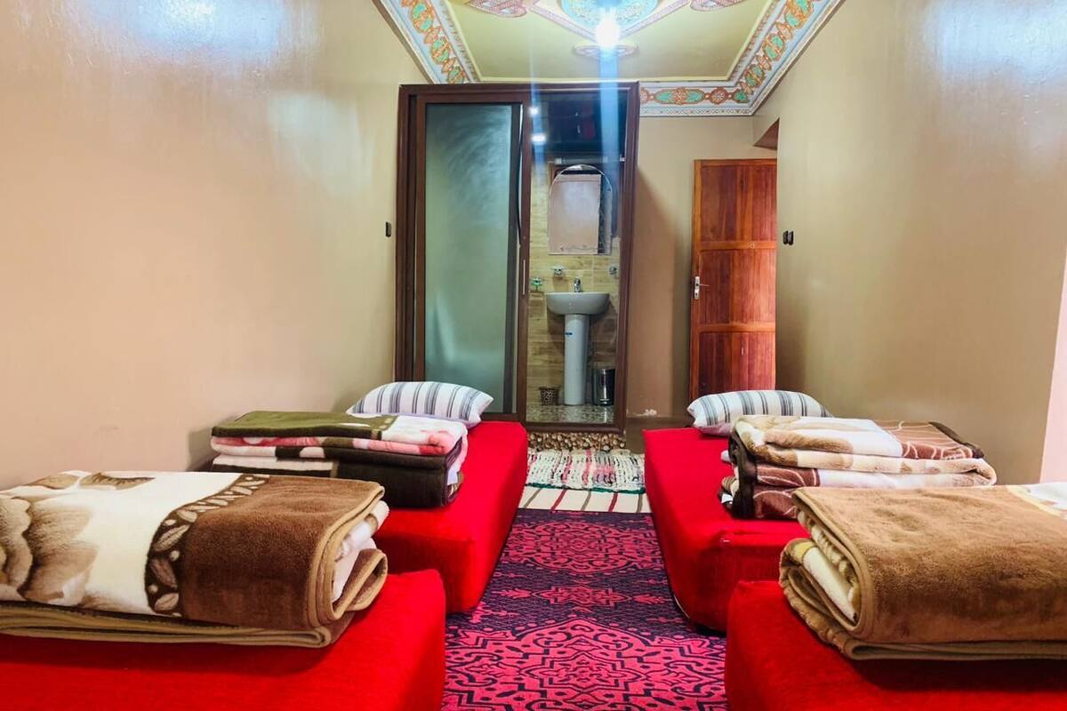 Una habitación con dos camas rojas, un tapete estampado y un baño visible a través de una puerta de cristal.