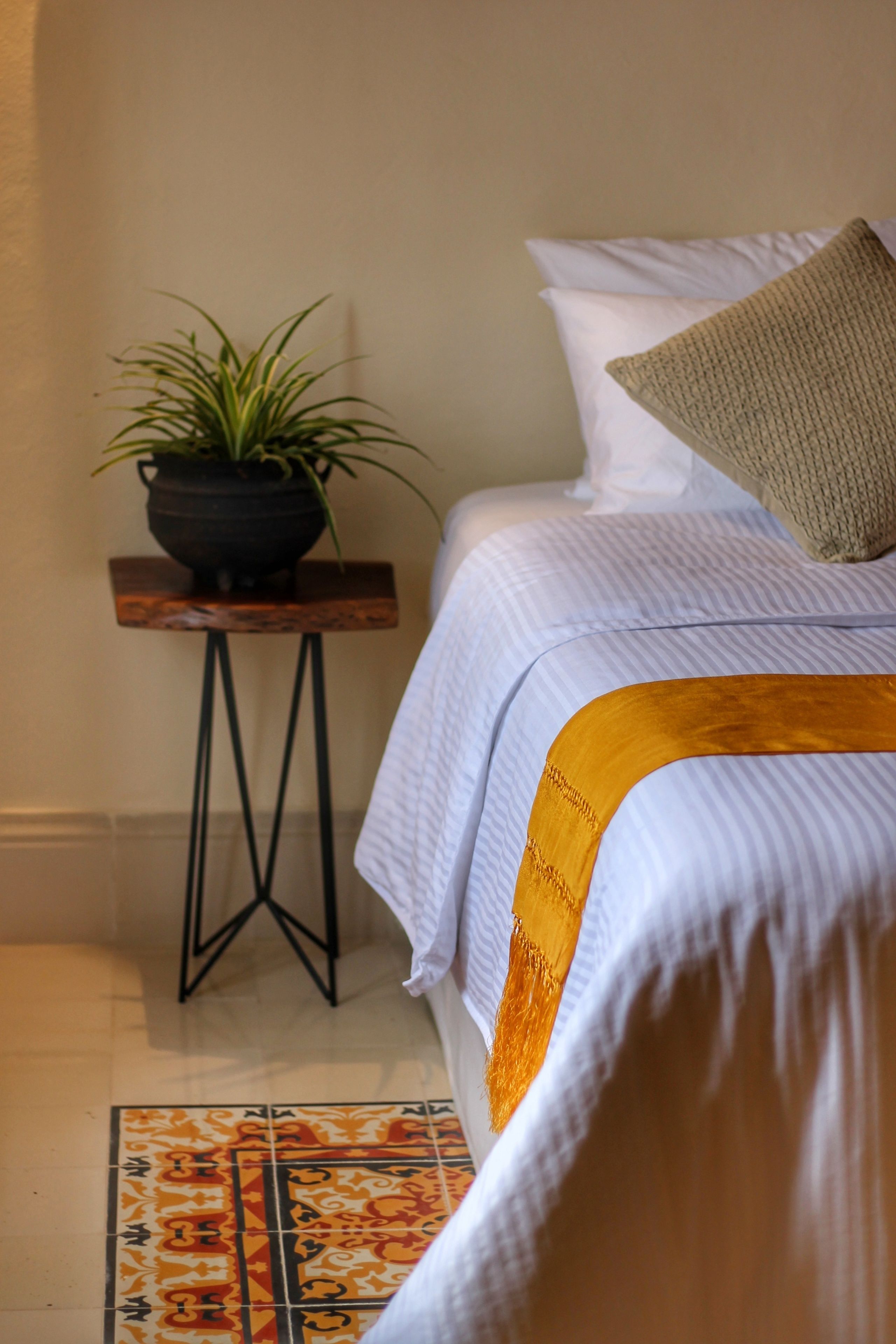 Una cama bien hecha con una colcha blanca y dorada, una mesita de noche de madera con una planta en maceta, y una alfombra estampada en el suelo.