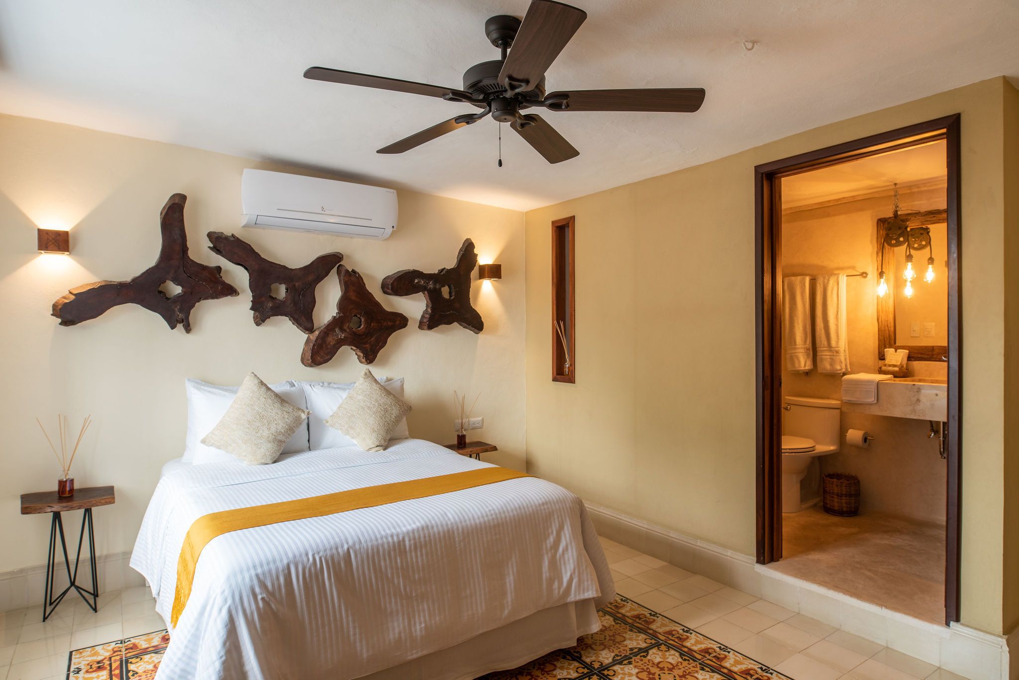 Une chambre d’hôtel avec un lit, un ventilateur de plafond, des éléments de décoration fixés au mur et une porte ouverte donnant sur une salle de bain.