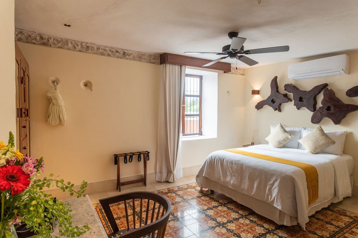 A bedroom with a bed, a ceiling fan, a window with curtains, a wall-mounted air conditioner, and a decorative wall piece.