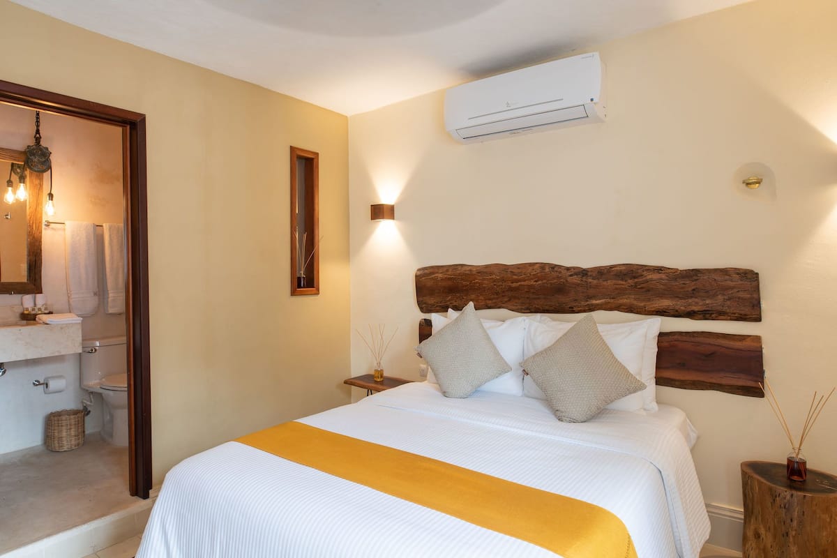 A bedroom with a wooden headboard, a bed with white bedding and a yellow accent, a nightstand with a vase, and an air conditioning unit on the wall.