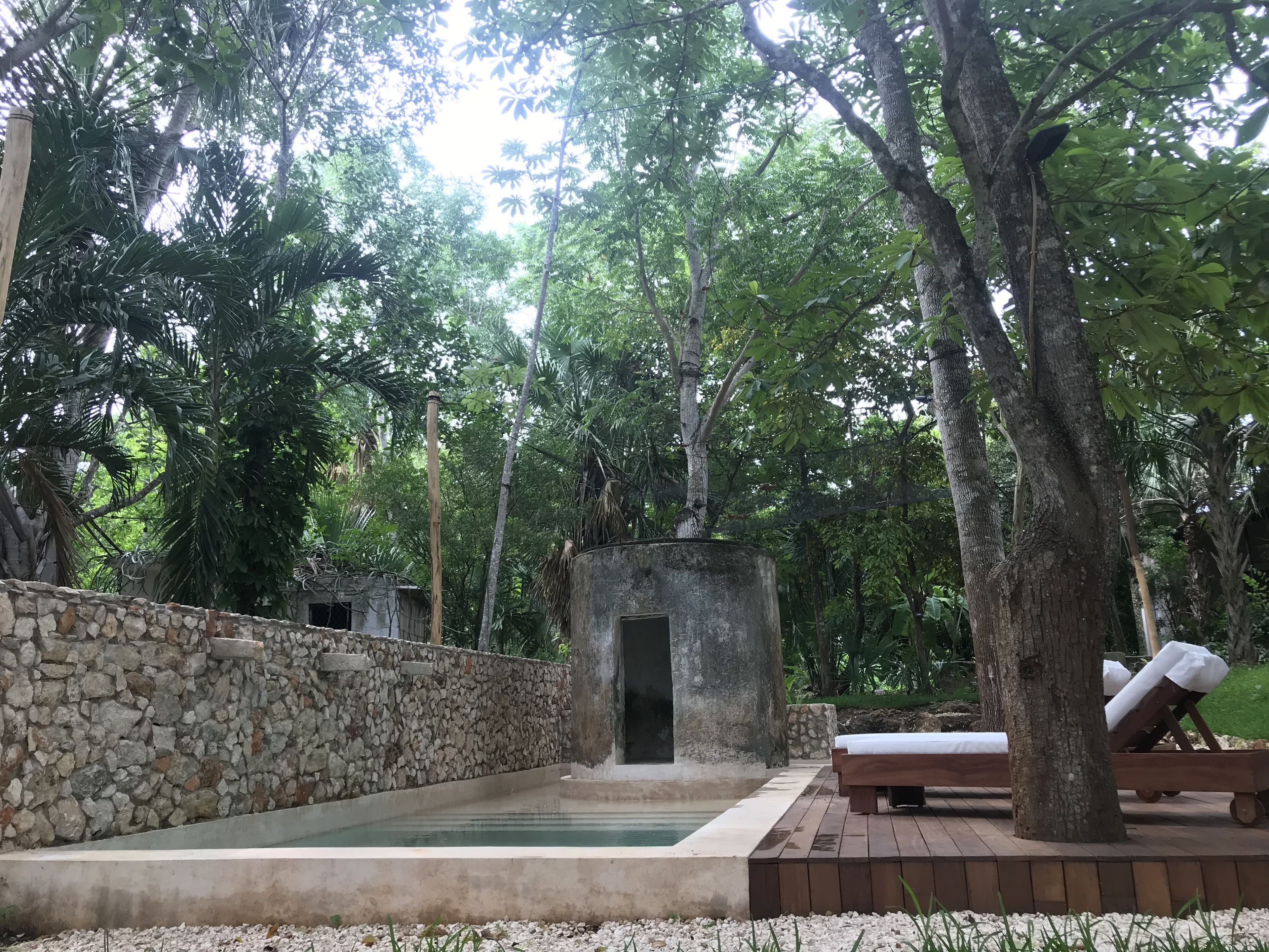 A serene outdoor area with a stone wall, a small pool, and wooden loungers under trees.