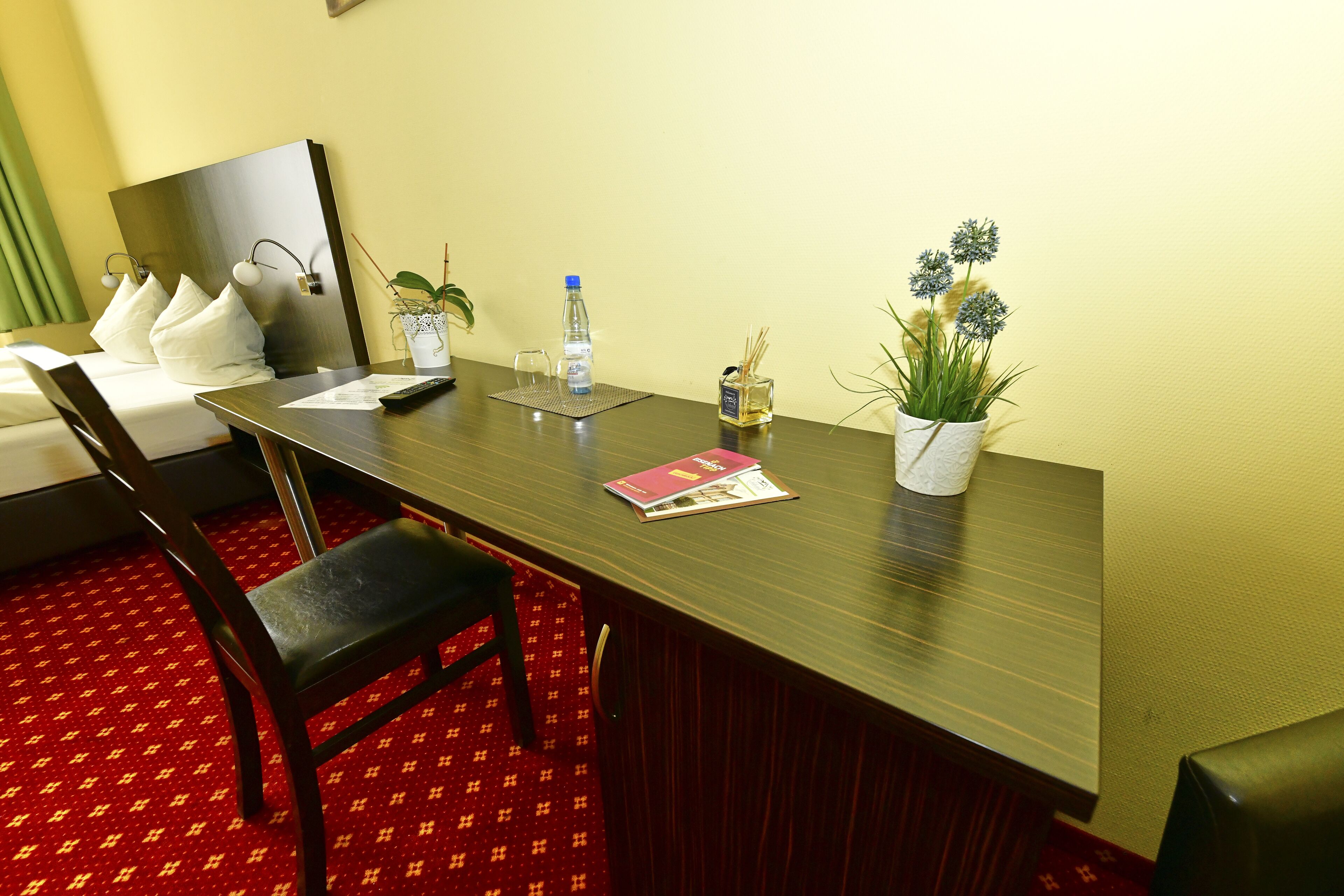 Habitación de hotel con una mesa de comedor de madera, dos sillas, una cama con almohadas blancas, una pequeña planta en maceta, una botella de agua y una revista.