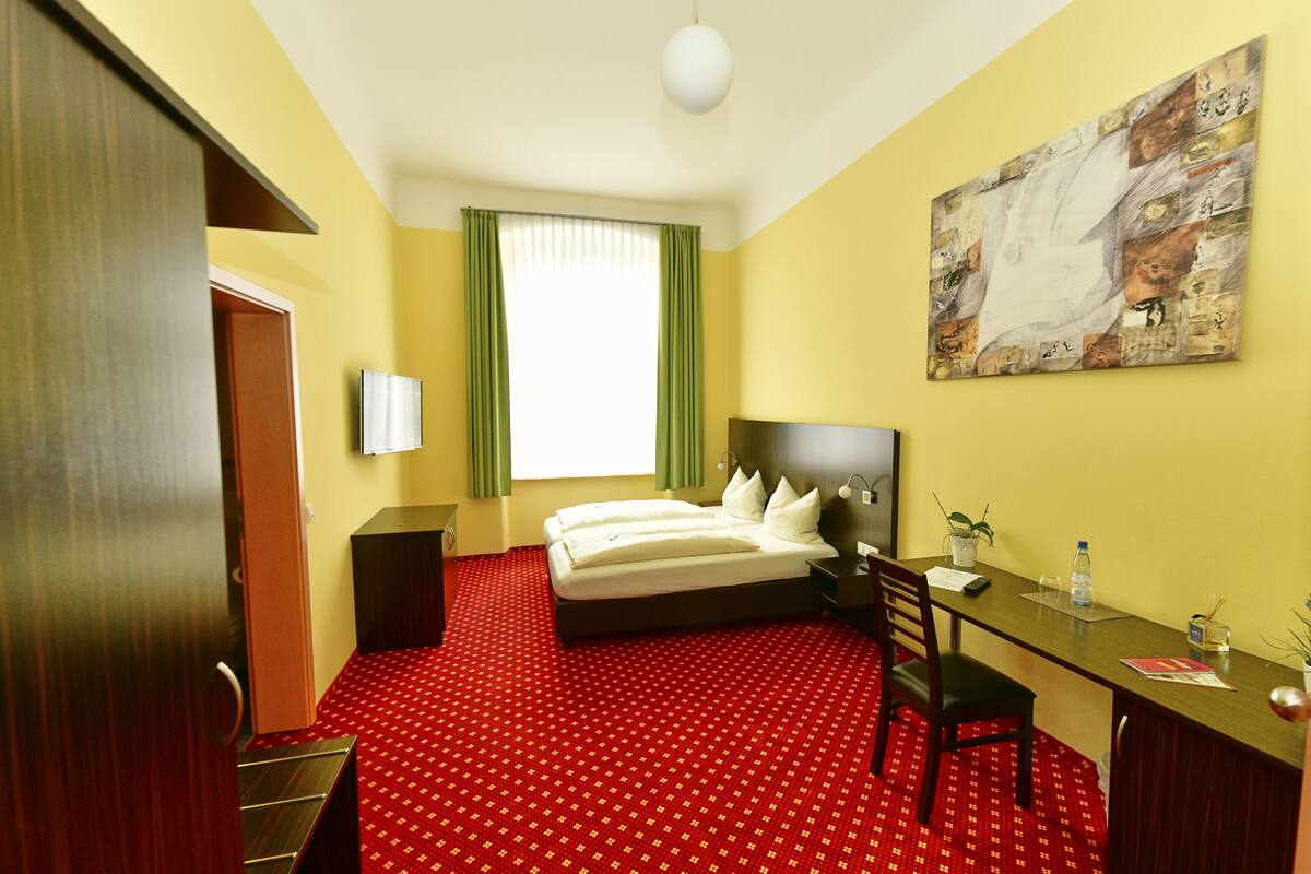 A hotel room with a bed, desk, chair, and a painting on the wall.
