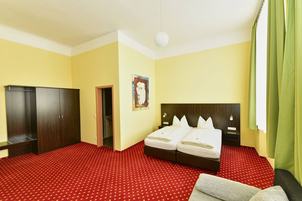 A hotel room with a red patterned carpet, a double bed, a wardrobe, and a green curtain.