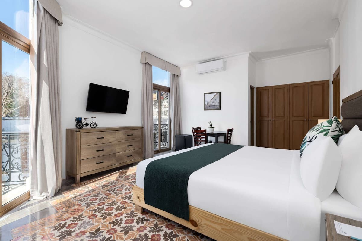 A hotel room with a bed, a TV, a wooden dresser, a desk with chairs, and a balcony with a view.