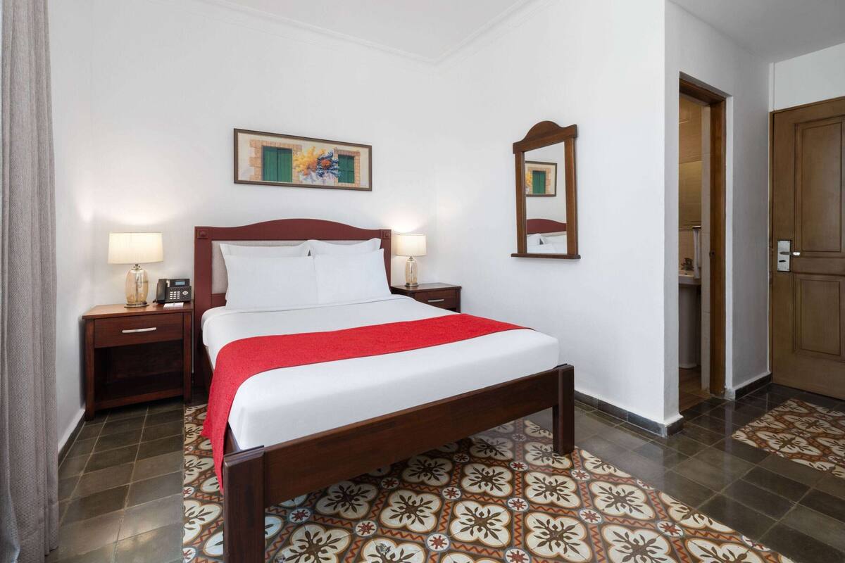 A hotel room with a wooden bed, a red throw, a nightstand with a lamp, a painting on the wall, and a mirror.