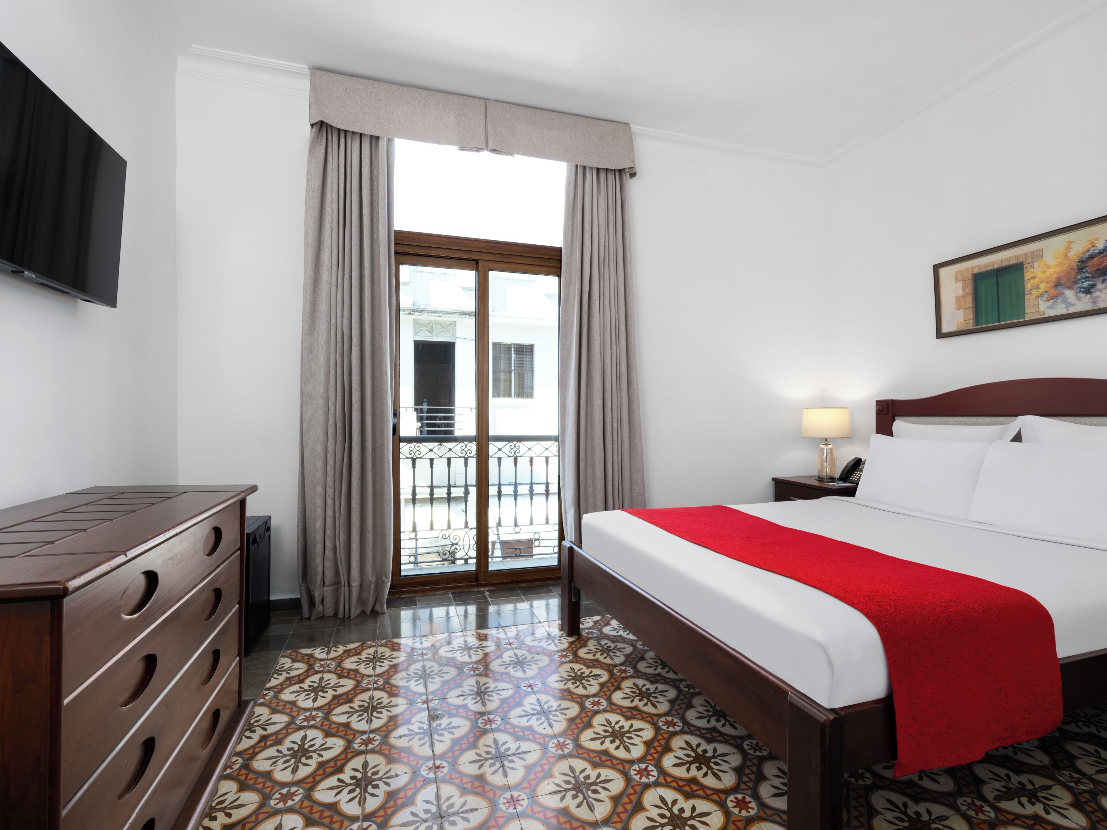 A hotel room with a bed, a dresser, a TV, a balcony, and a painting on the wall.