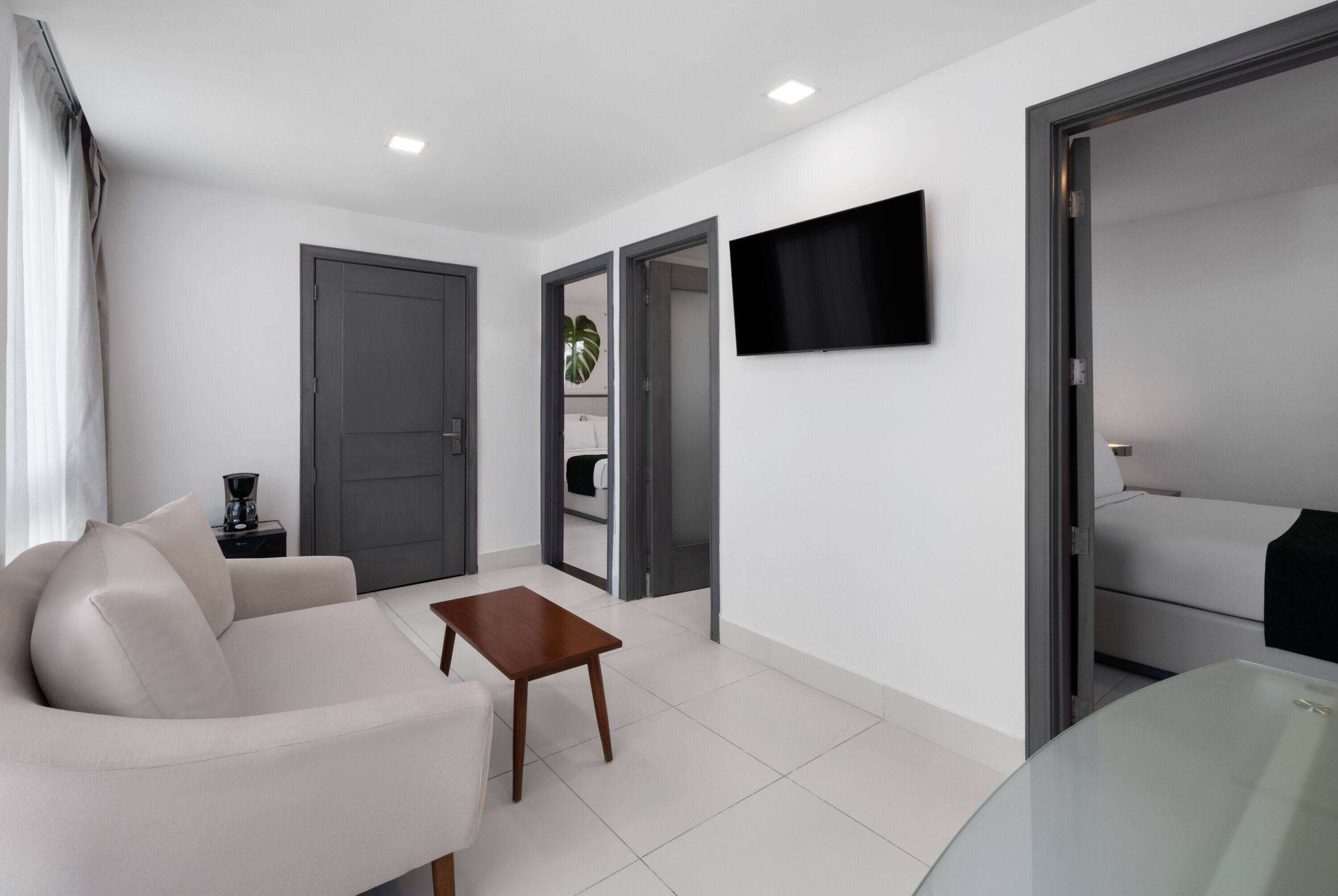 Een moderne woonkamer met een bank, een salontafel, een aan de muur bevestigde televisie en een deur naar een andere kamer.