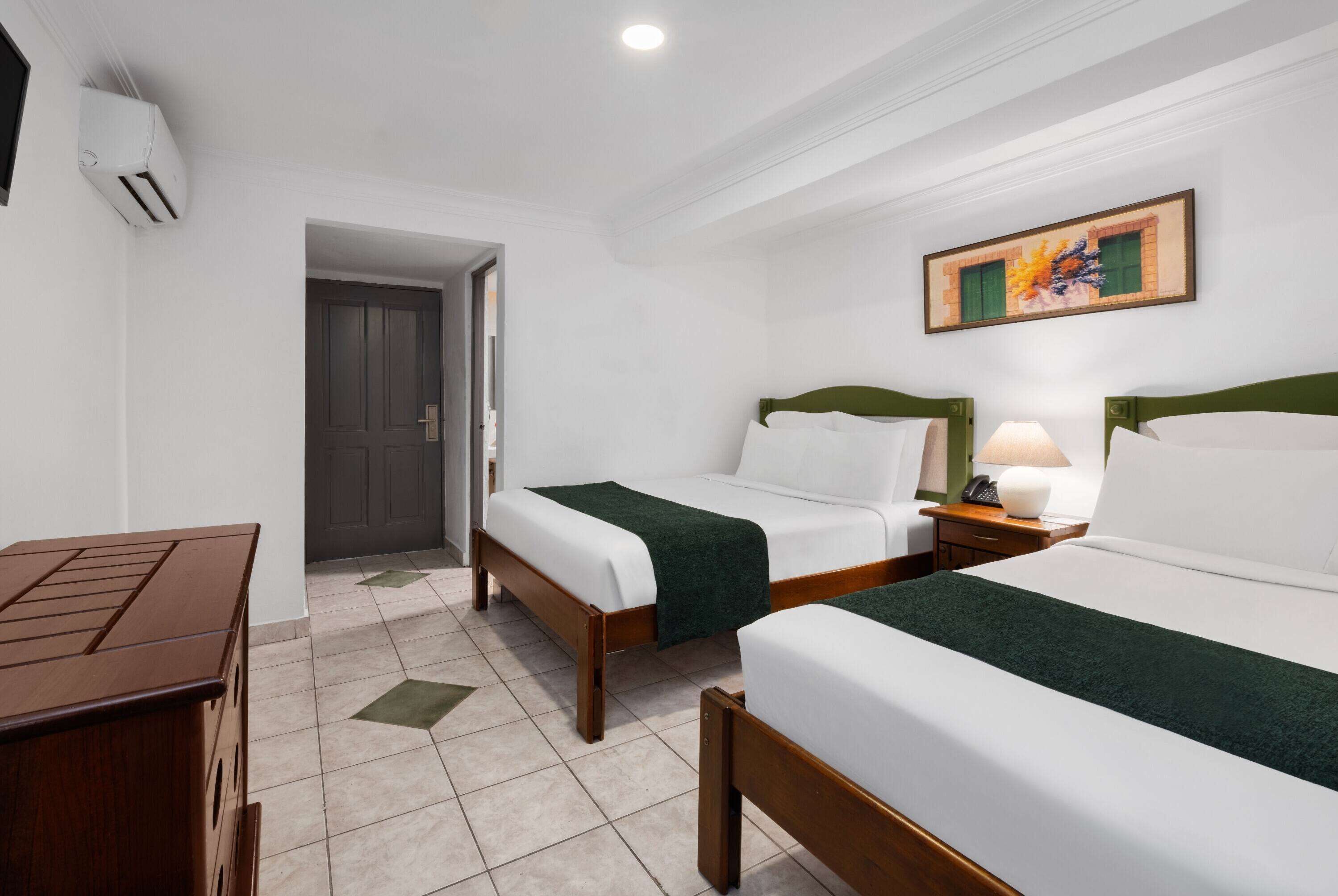 Une chambre d’hôtel avec deux lits simples, une commode en bois, un bureau et un tableau au mur.