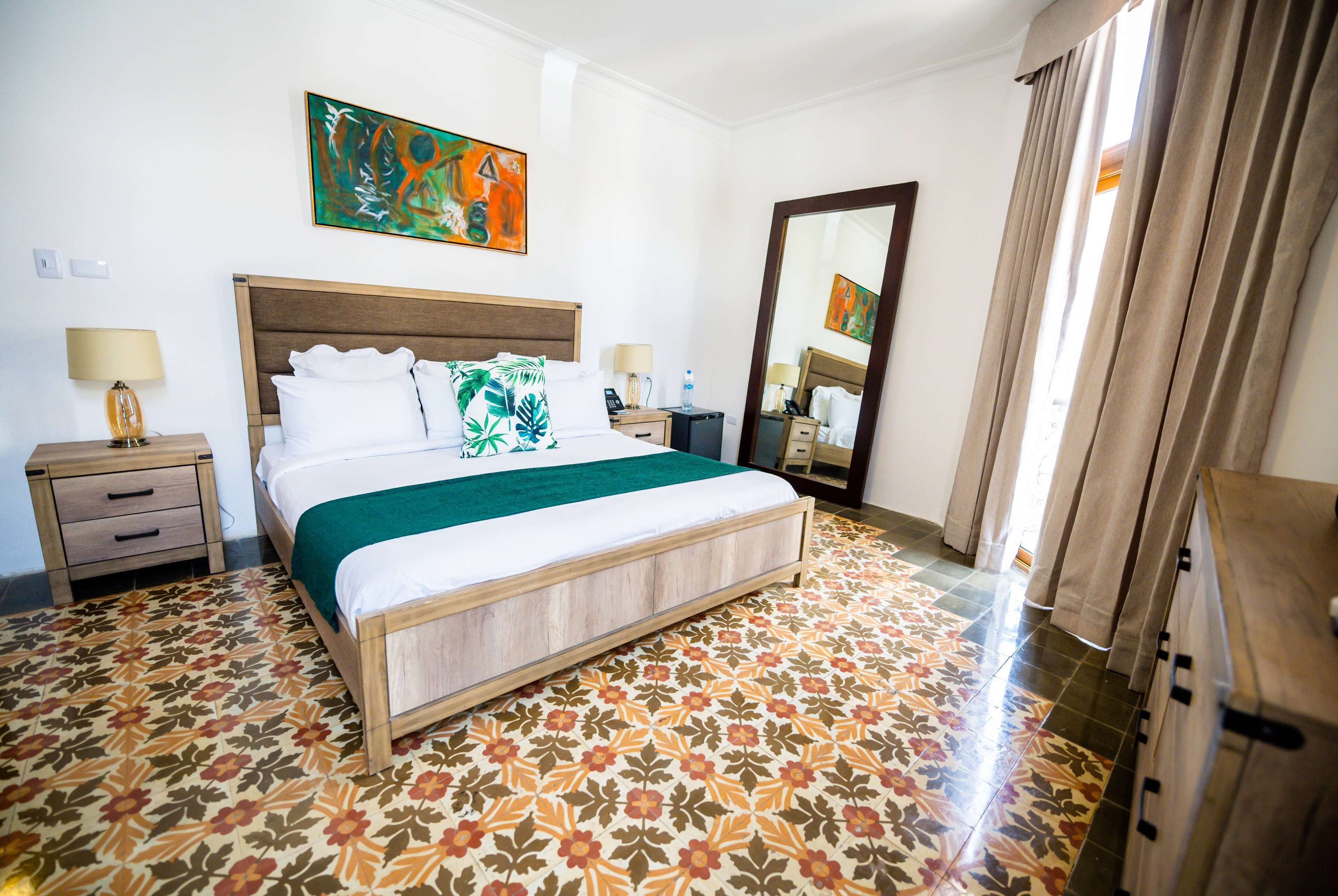A hotel room with a bed, two bedside tables, a nightstand, a mirror, and a window with curtains.