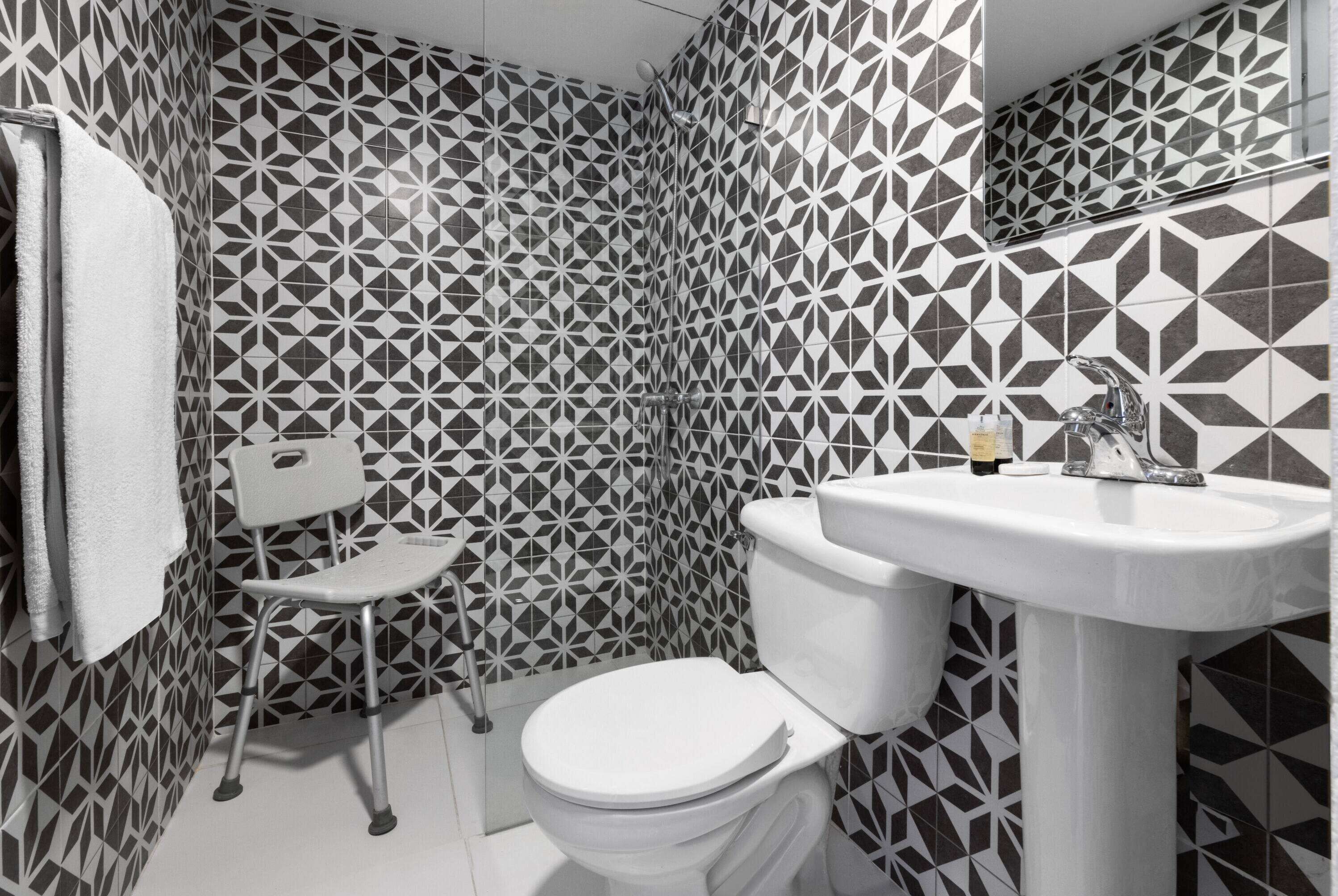 Un baño con pared de diseño geométrico, inodoro blanco, lavamanos y una pequeña silla blanca.