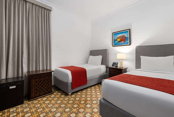 A hotel room with two beds, a small table, a lamp, a painting on the wall, and a patterned floor.
