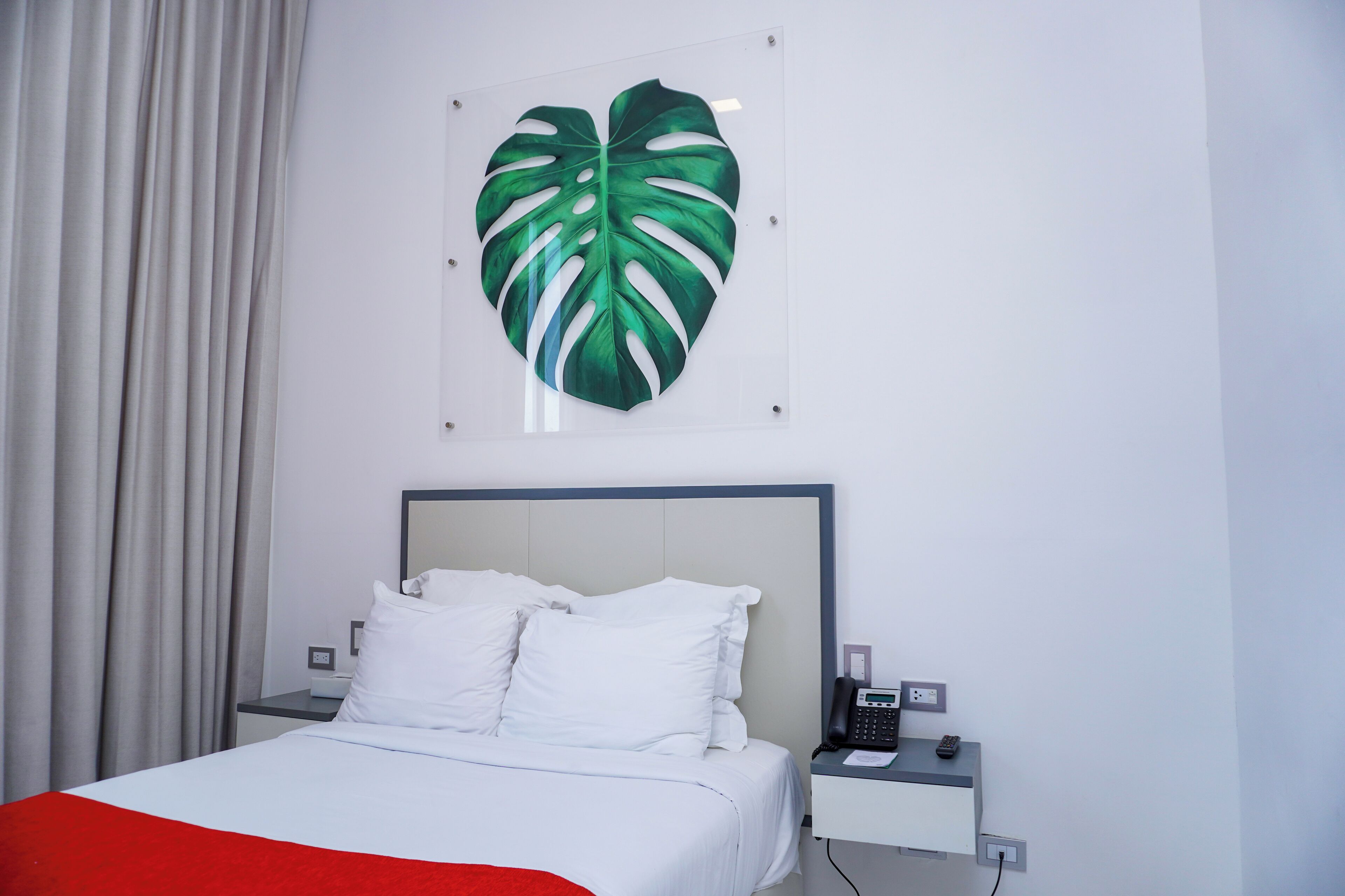 A hotel room with a bed, white bedding, a nightstand with a phone, and a large green leaf artwork on the wall.