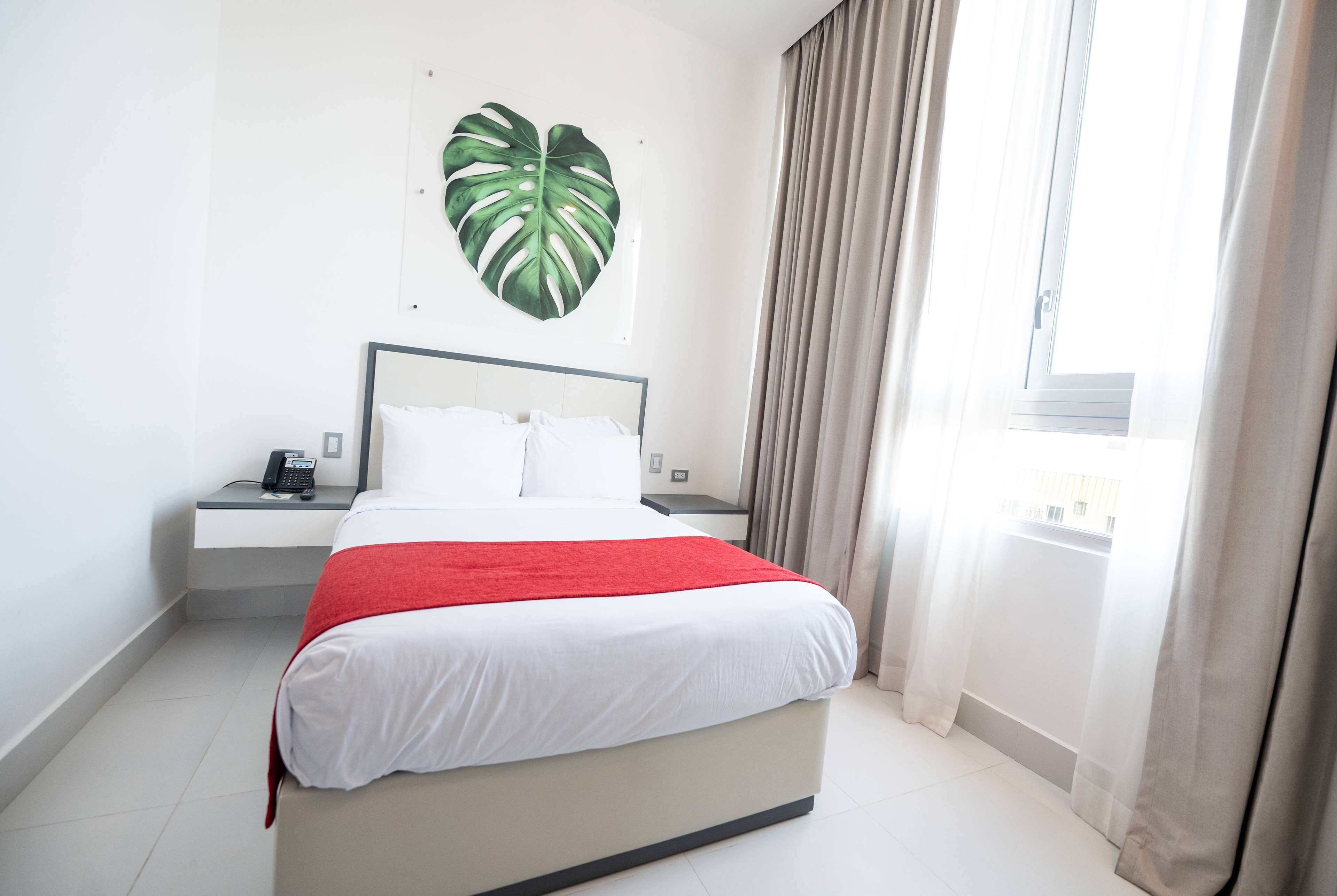 A hotel room with a bed, a bedside table with a phone, a heart-shaped wall decoration, and a window with curtains.