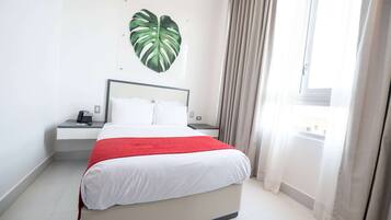 A hotel room with a bed, a bedside table with a phone, a heart-shaped wall decoration, and a window with curtains.