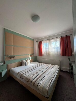 A bedroom with a bed, a nightstand, a dresser, a window with curtains, and a radiator. - Hotel Route 84 (Bad Langensalza)