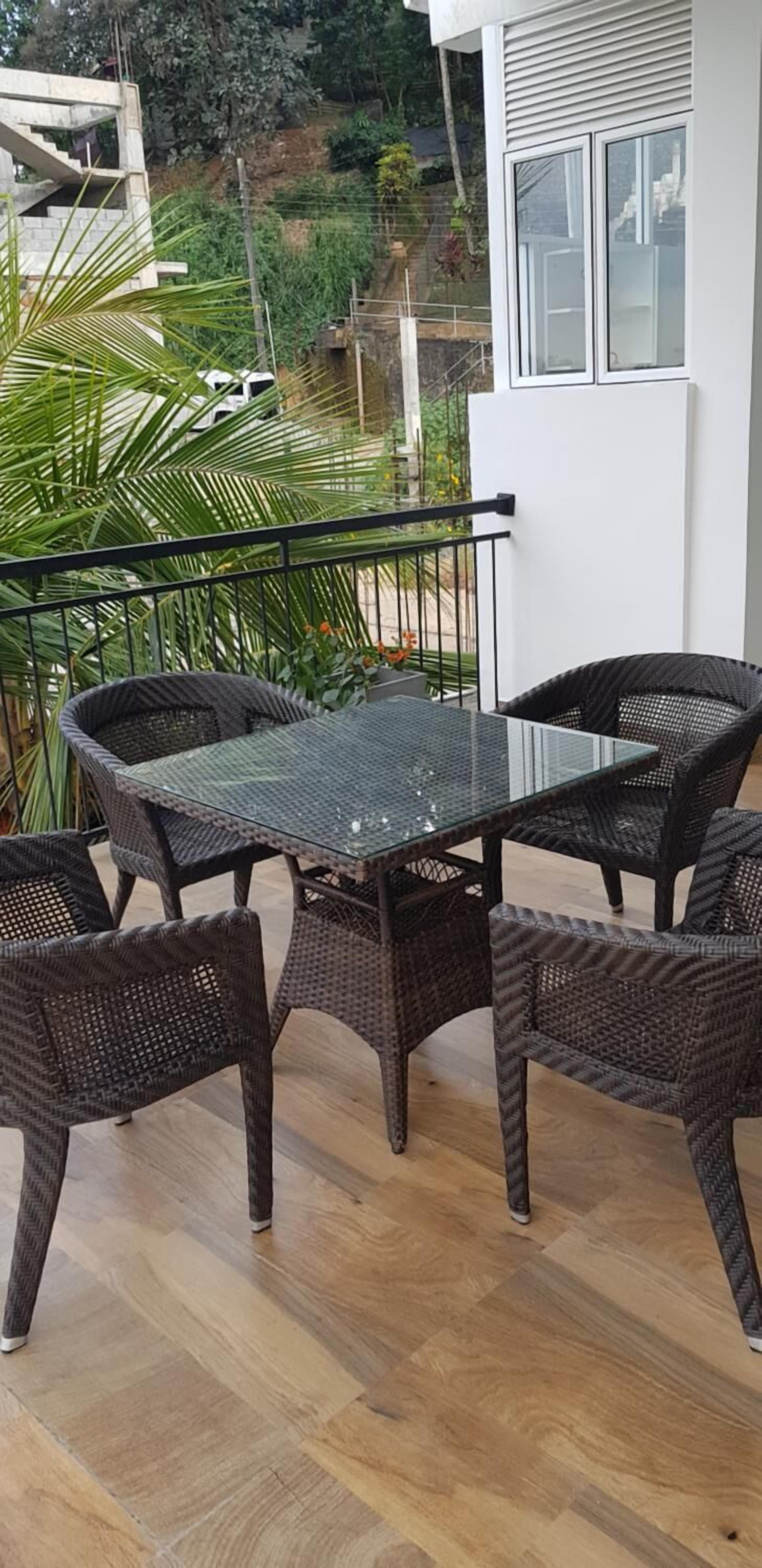 Un balcon doté de meubles en osier et d’une table en verre, offrant une vue sur un jardin et un bâtiment blanc.