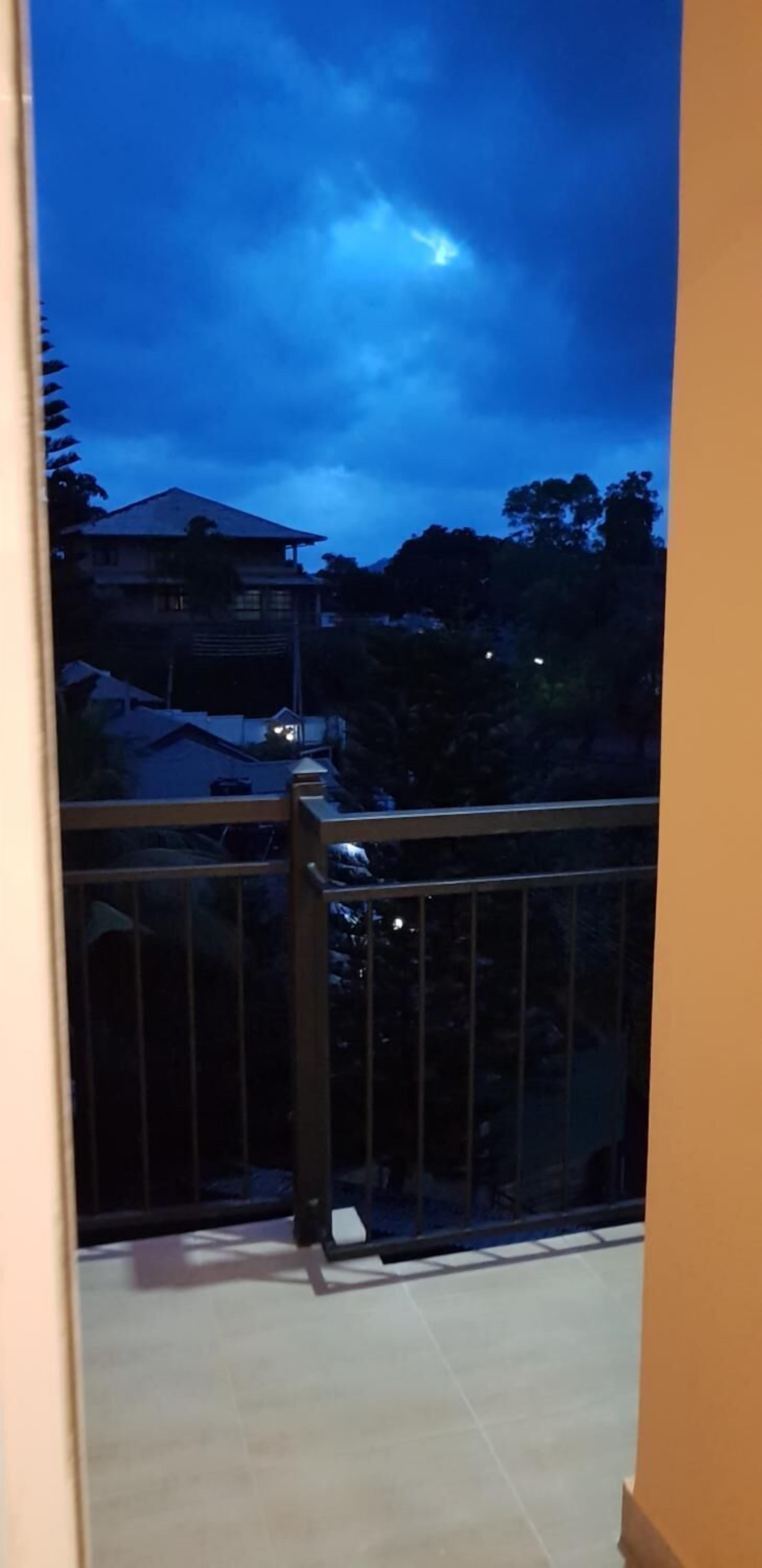 Un balcon doté d’une rambarde en métal, d’un sol carrelé et offrant une vue sur une maison et des arbres, le tout sous un ciel sombre.
