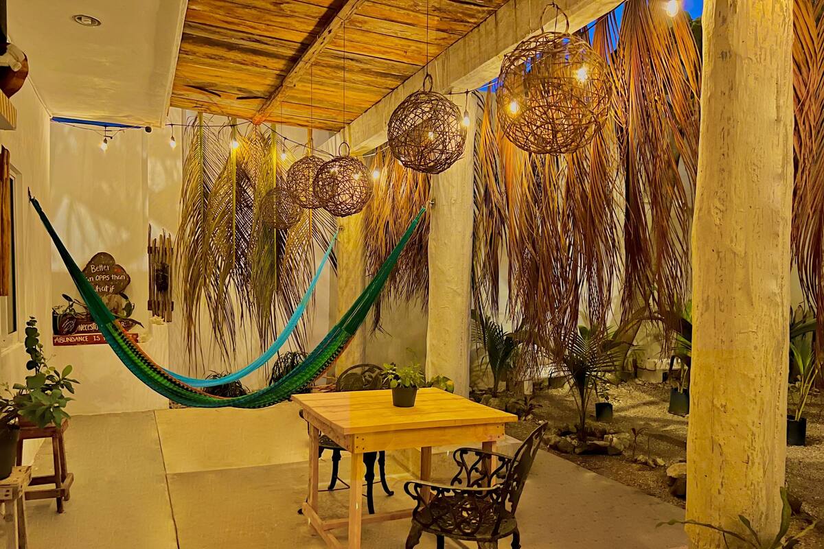 Un espace de restauration extérieur accueillant, avec une table en bois et des chaises en métal, entouré de luminaires suspendus en fibres tressées et d’un hamac.