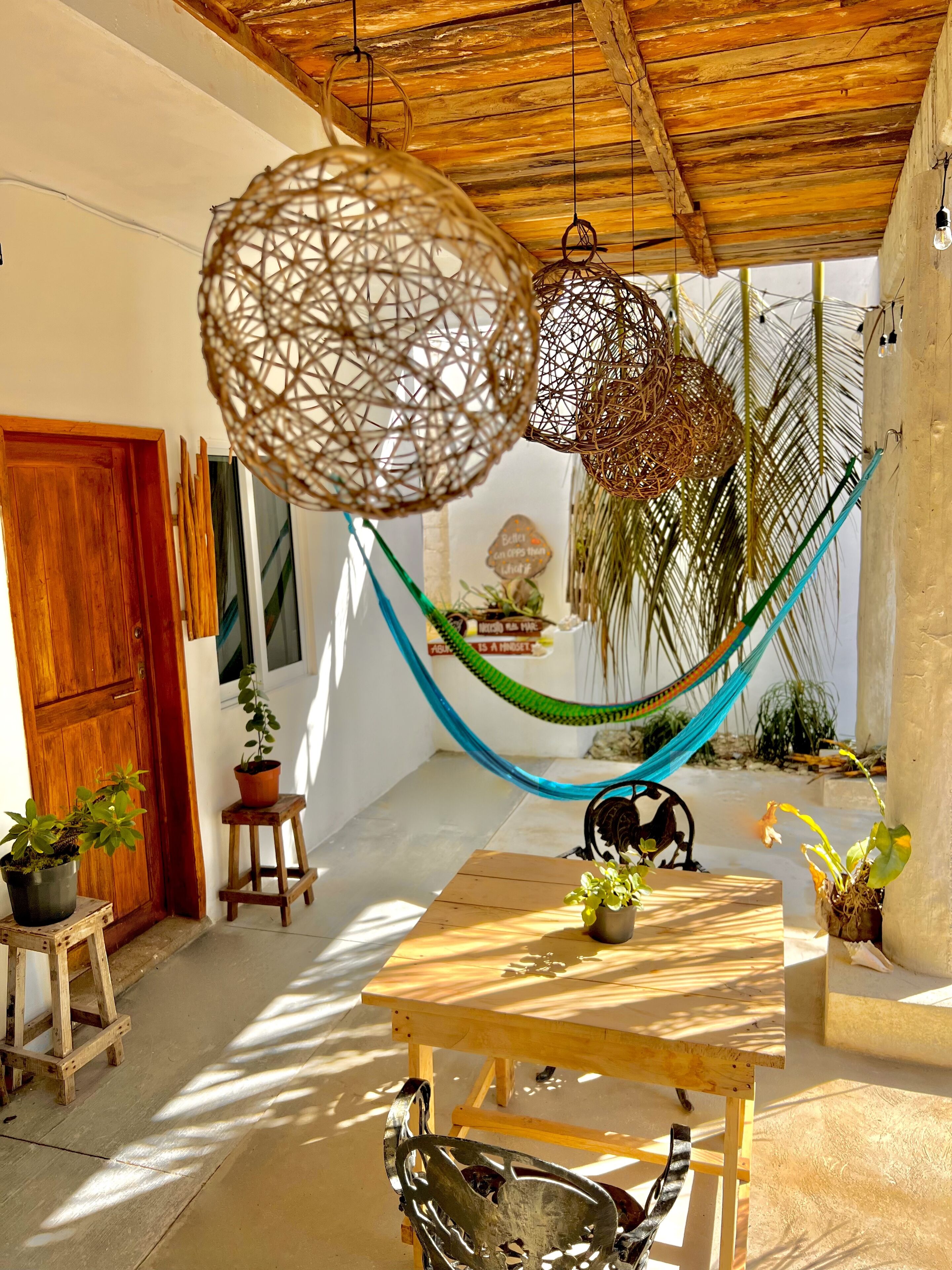 Una mesa de madera con plantas en macetas, una hamaca y lámparas colgantes de tejido en un espacio exterior iluminado por el sol.