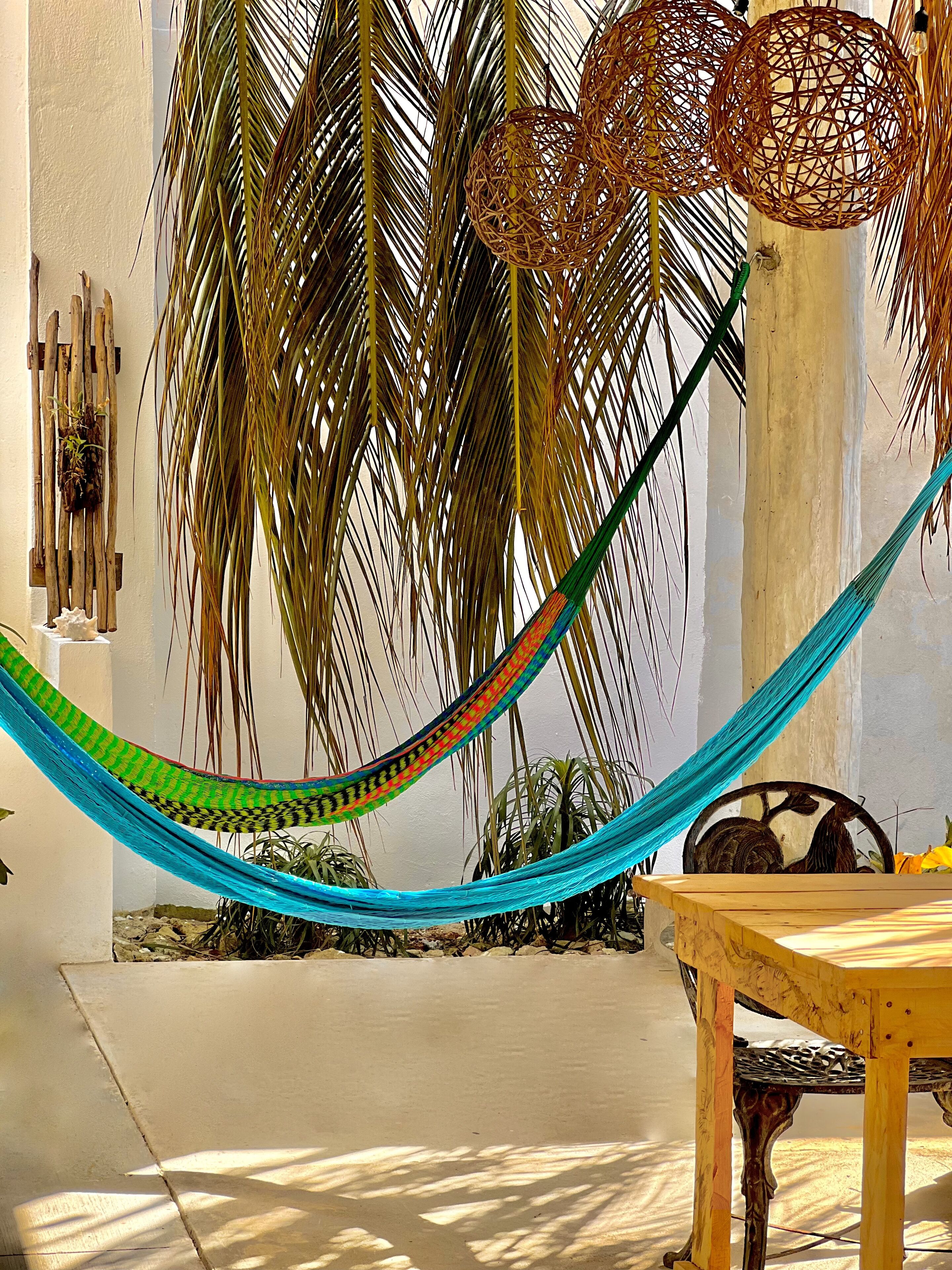 A hammock tied between palm trees, a wooden table, and a rustic chair in a sunny outdoor setting.