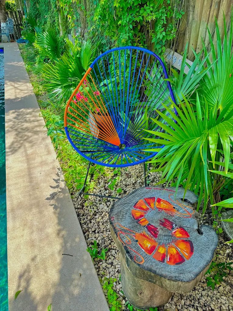 A garden with a blue chair, a wooden table with painted designs, and a pool.