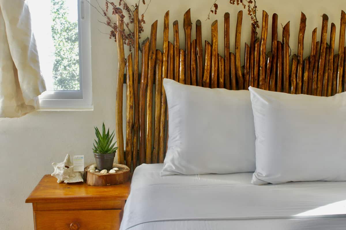 Un lit avec une literie blanche, une table de chevet en bois ornée d’un pot de plantes, un mur décoratif et une fenêtre donnant sur des arbres.