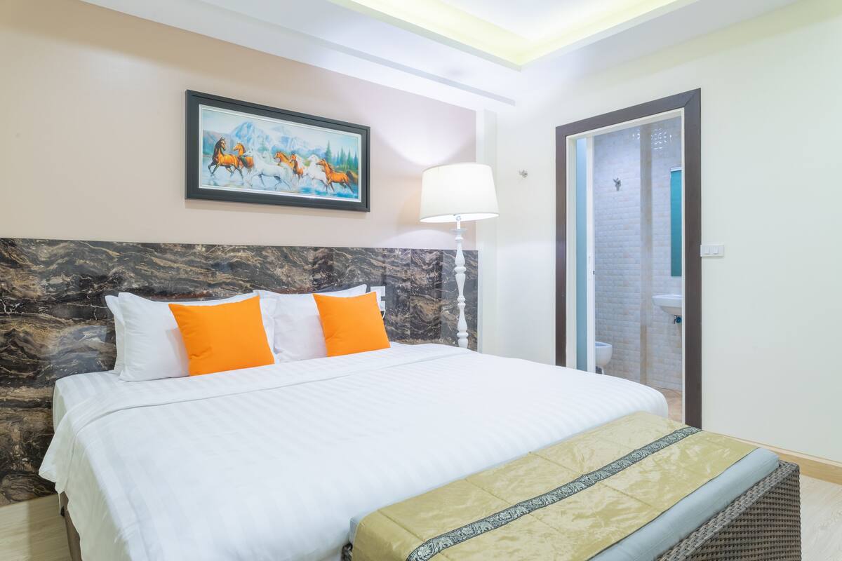 A hotel room with a bed, a painting, a lamp, and a bathroom visible through an open door.
