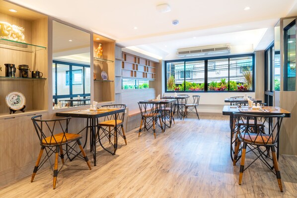 A modern dining area with wooden flooring, black metal chairs, and tables. There are shelves with decorative items and a large window with a view of greenery.