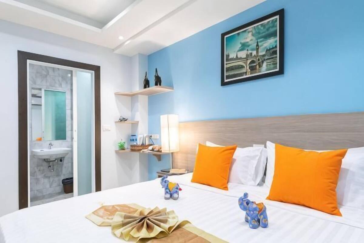 A hotel room with a bed, a nightstand, a lamp, a painting, and a bathroom with a sink and mirror.