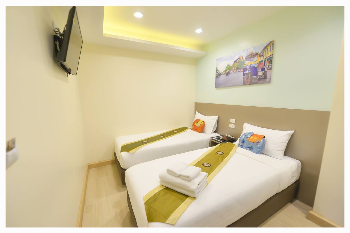 A hotel room with two beds, each with a green and white bedspread, a flat-screen TV mounted on the wall, and a painting of a street scene above the beds.