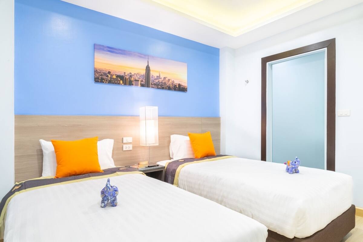 A hotel room with two beds, a cityscape painting on the wall, a nightstand, and a door.
