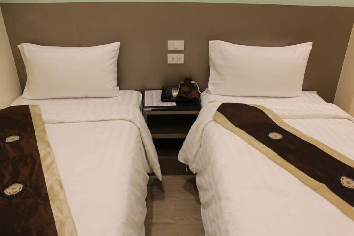 A hotel room with two single beds, a nightstand with a lamp, and a wall socket.