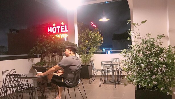 Un uomo seduto a un tavolo con sedie, un vaso di fiori e, sullo sfondo, un neon con la scritta 